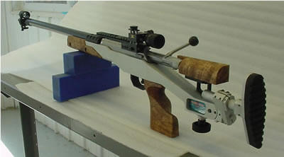 Rifle on a white table with silver body, black scope, and brown wooden grip.
