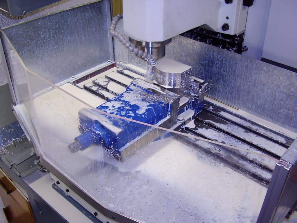 White and blue CNC machine with water coolant system, surrounded by transparent plastic cover, featuring flat surface with foam-like material.