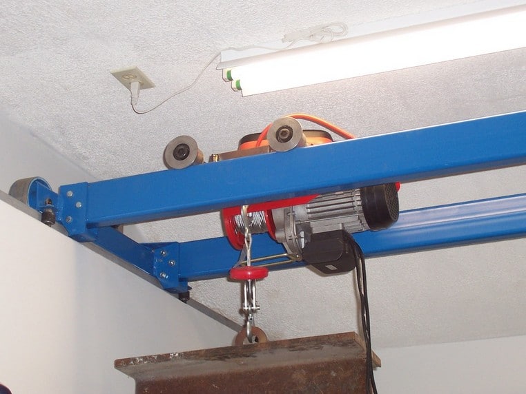 Blue metal beam with red and black components suspended from ceiling, featuring two wheels and a red winch.