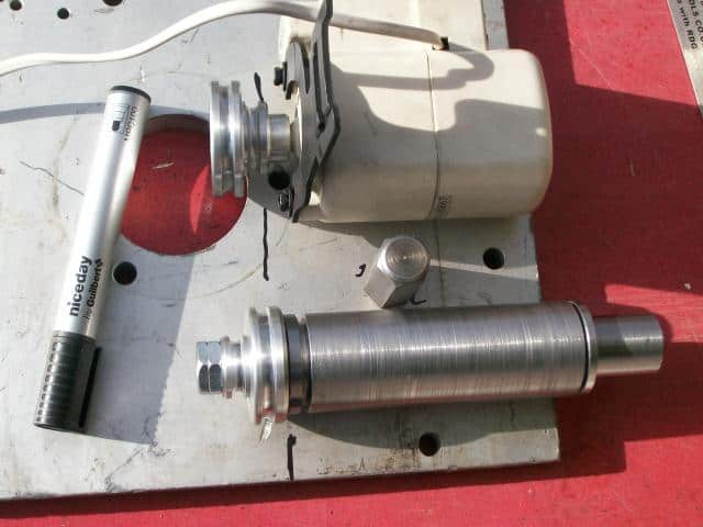 Metal parts and tools on a white surface with red undertones, featuring a threaded cylinder and small metal pin.
