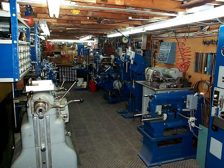 Well-organized machine shop interior with blue machinery and tools on a wood-planked ceiling and gray-carpeted floor.