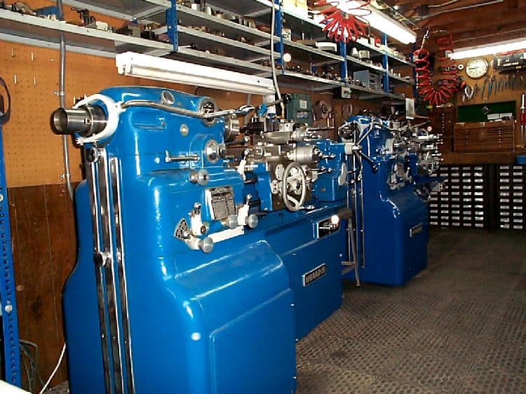 Large blue metalworking lathes in a workshop setting with metal components and wooden walls.