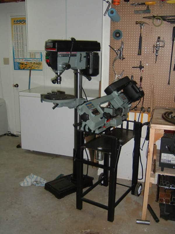 Cluttered workshop with drill press and bandsaw on concrete floor and surrounding tools.