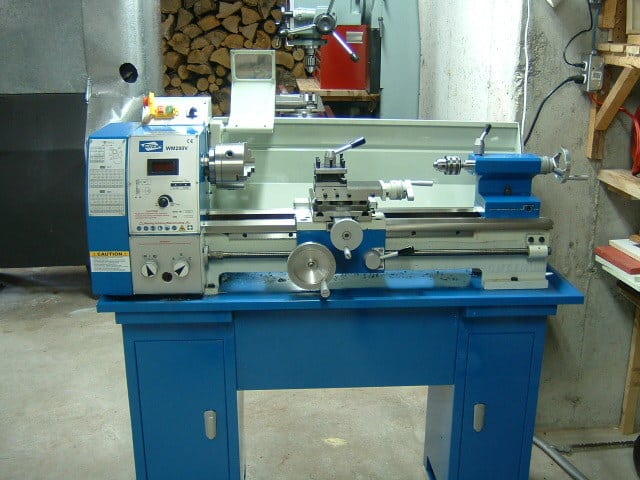 Blue and silver lathe on blue cabinet in well-lit room with gray walls and woodpile in background.
