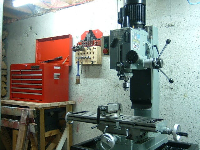 Compact workshop setup with red toolbox, pegboard, and gray metal drill press on wooden workbench against white concrete wall.