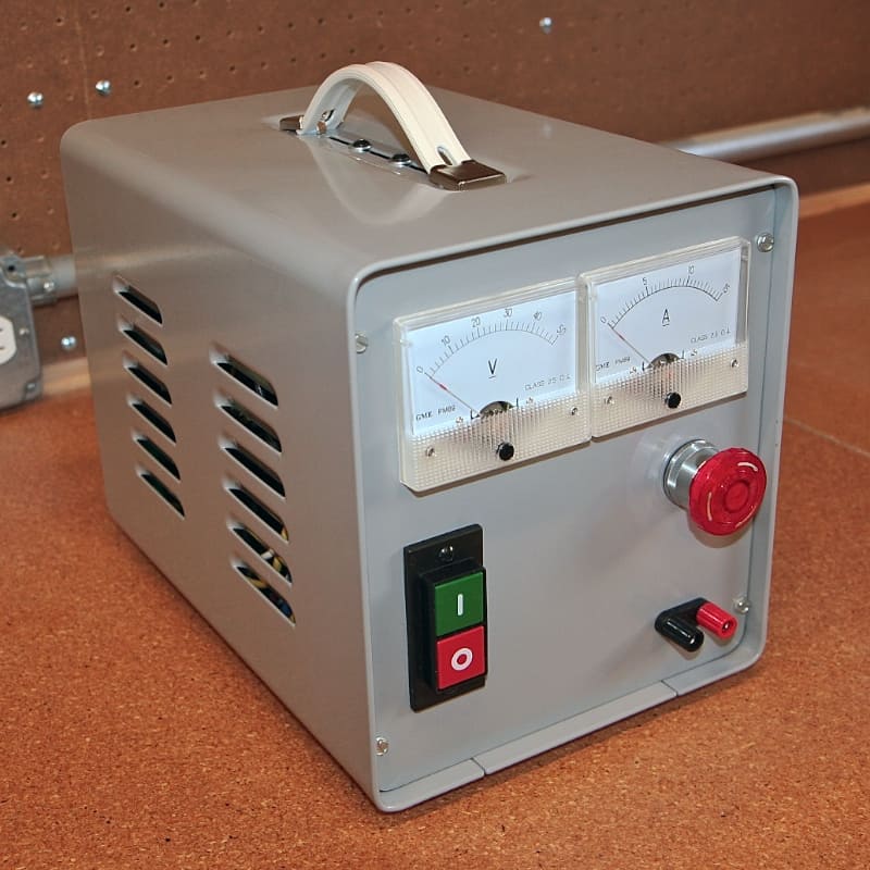Metal gray electrical box with dials, red button, and strap handle on a brown surface.