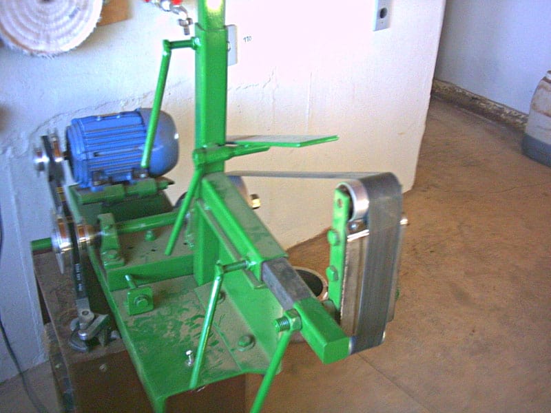Green metal device with blue motor and attachments on brown floor in front of white wall.