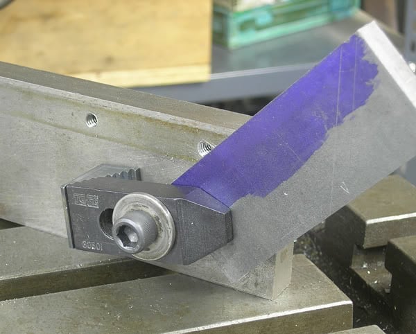 Metal component with purple tape strip on flat surface featuring drilled holes, silver-colored material, and background components in various states of completion.