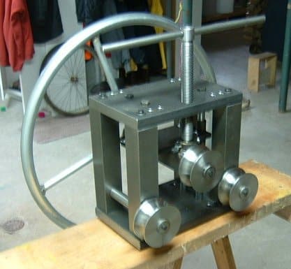 Stainless steel tube bender with adjustable handle on wooden bench in workshop setting.