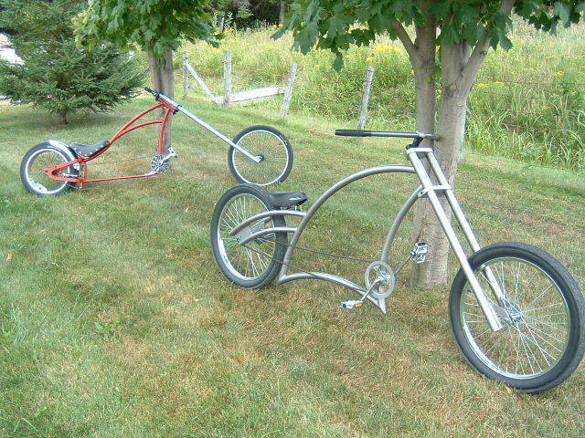 Two custom bicycles, one silver and black, the other red and white, parked under a tree in a grassy area.