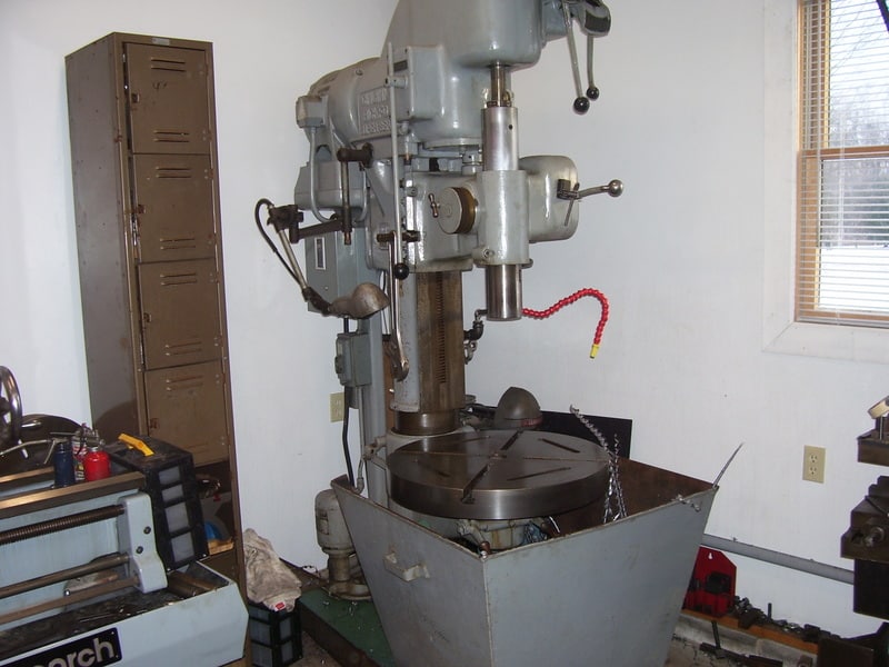 Large industrial drill press machine in cluttered workshop setting.
