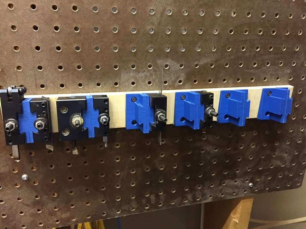 A 3D-printed rack with six blue and black QCP holders mounted on a pegboard in a workshop or machine shop setting.