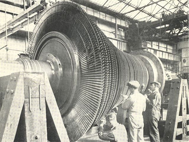 Industrial workers examining a large cylindrical machine with ridged sides.