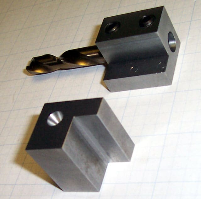 Two gray metal objects with square bases on a white surface with grid lines: a twisted drill bit holder and a corresponding drill bit.