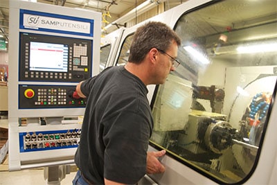 Man in black t-shirt standing beside window looking at machinery and computer screen in a machine shop.