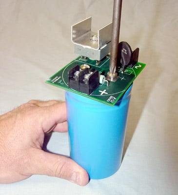 A person's hand holds a blue cylinder with a green electronic circuit and metal components on top.