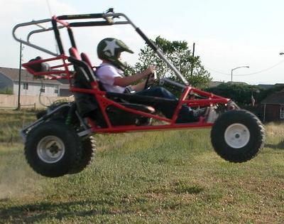 A red go-kart with a black roll cage and a person wearing a white star patterned helmet on a grassy field with houses and trees in the background.