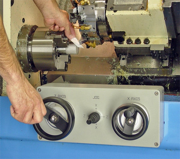 A man's hand grasps a metal piece on an industrial machine with control dials, in a workshop or factory setting.