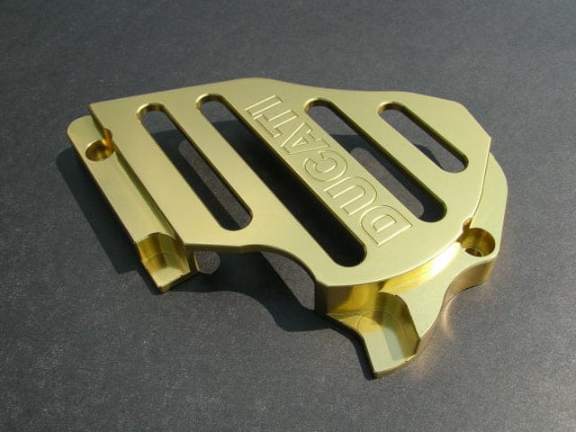 A shiny gold metal part with four horizontal slots and embossed text "DUCATI" on a dark gray surface.