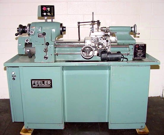 Light blue Feeler HLV-H Clone lathe with various controls and levers, against a white wall on a dark floor.