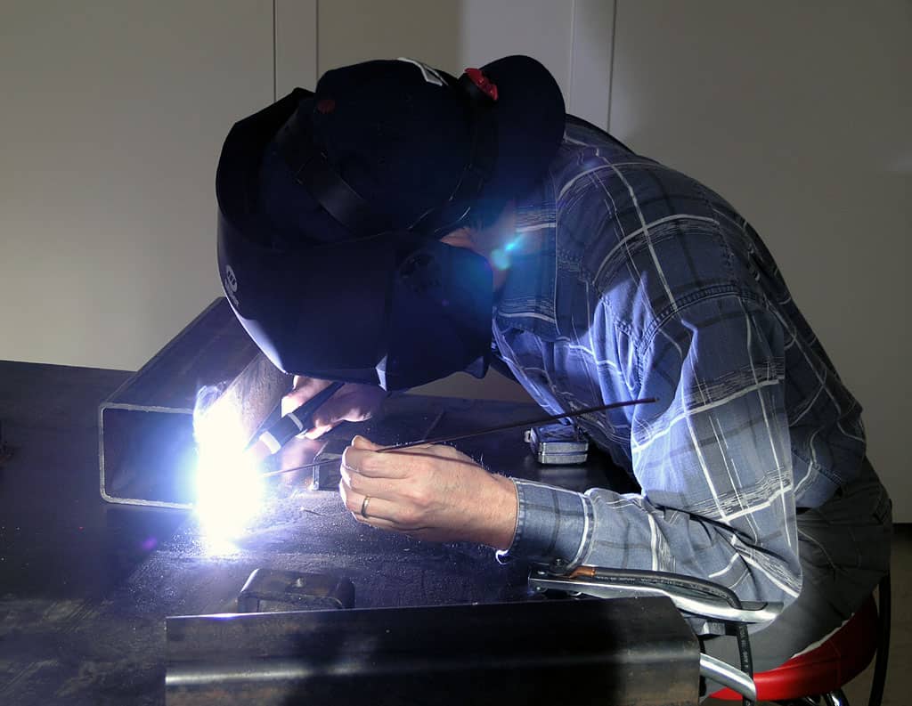 Man in plaid shirt and welding helmet focusing on metalwork with tool.