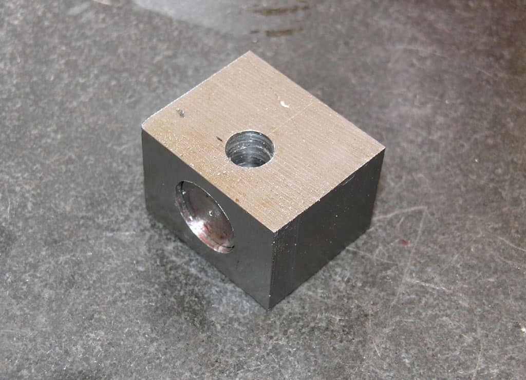 Small metal block with hole and circular cutouts, machined steel or aluminum construction.