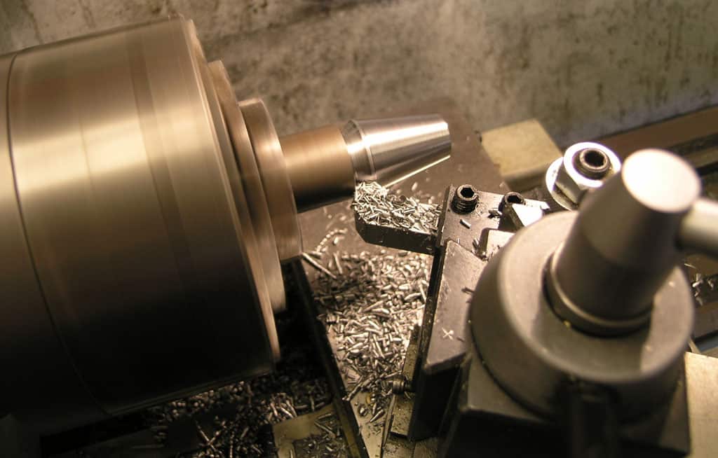 Metalworking lathe with cylindrical material being machined, tool rest and tailstock visible, shavings on the bed.