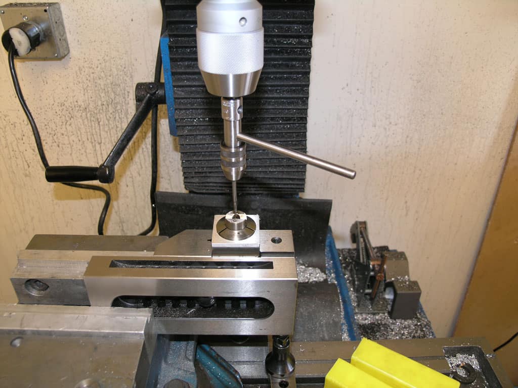 A drill press with a chuck holding a drill bit, set on a black rubber mat in a workshop or machining facility.