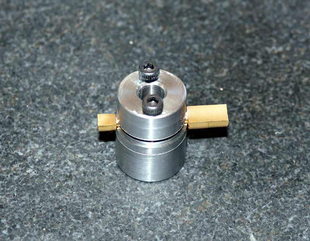 Small silver metal component with screws and cylindrical protrusions on dark gray surface.