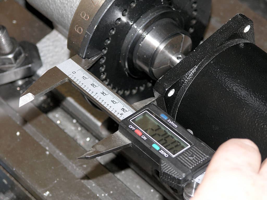 A black and silver digital caliper with open jaws next to a large metal object.