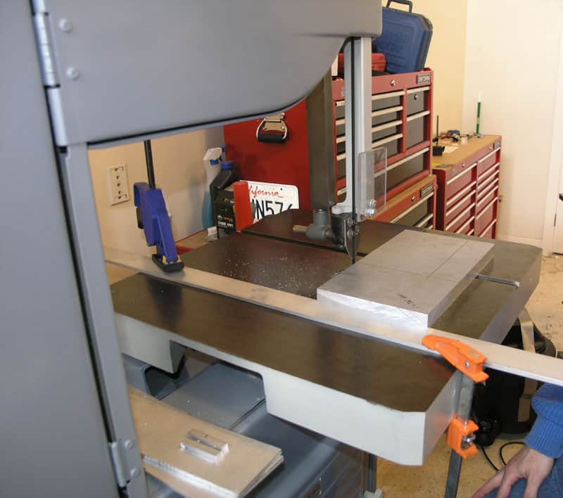 A bandsaw in a workshop setting with various metalworking tools and equipment visible in the background.