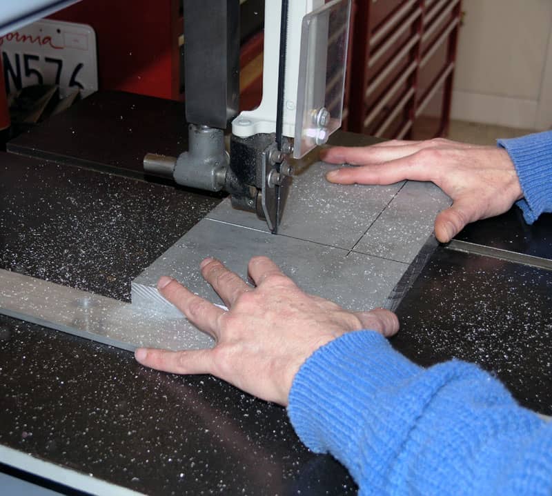 A person's hands operating a bandsaw on a black table in a workshop cutting through aluminum.
