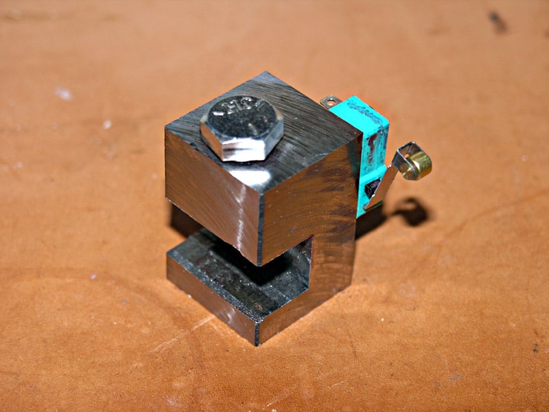 Small metal vise with square base and flat top, featuring a large bolt on the side, set on a brown surface.
