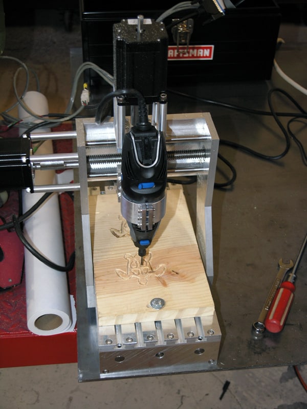 Custom-built milling machine on wooden base with drill bit, metal frame, and wires next to a black Craftsman toolbox.