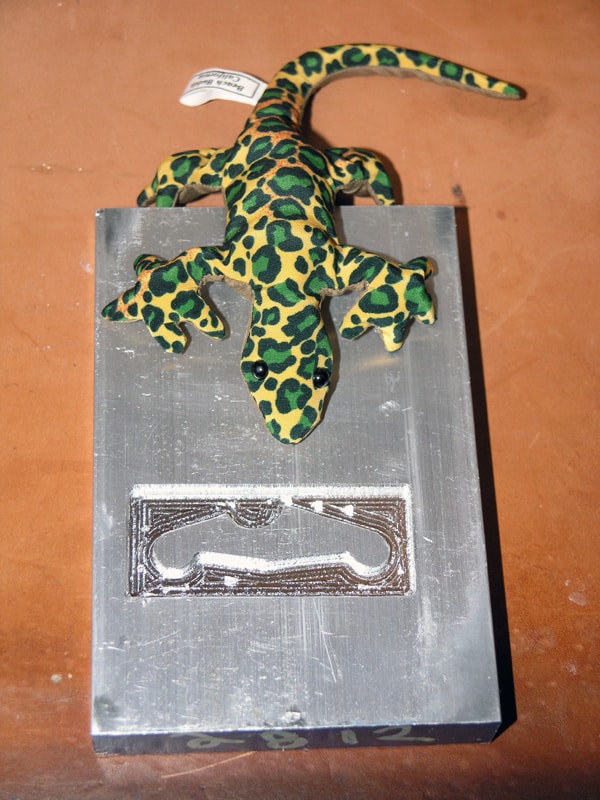 Small lizard figurine on brown tabletop with metal tool impression below.