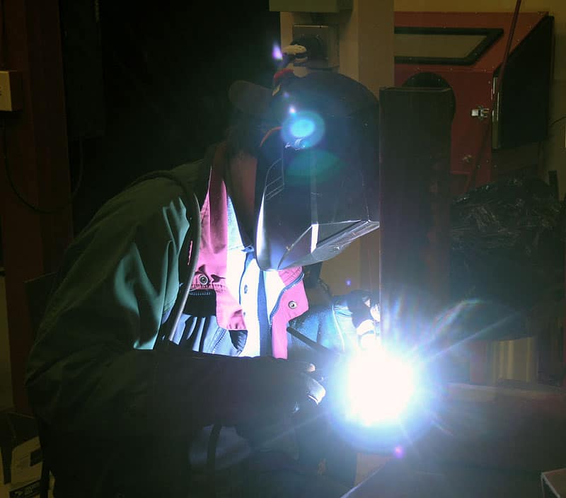 A person in green scrubs welding with a mask, holding a welding torch, in a workshop background.