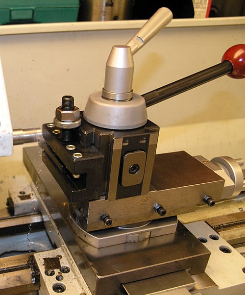 Metalworking lathe tool rest with lever-activated mechanism for adjusting tool position, featuring black base plate, silver arm, red ball, and silver bar mounted to metal lathe bed.