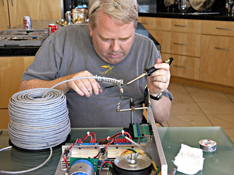 A man sitting at a workbench with soldering iron, wire, and tools.