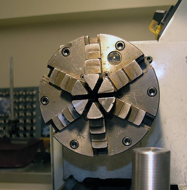 A metal collet chuck with radial grooves and evenly spaced holes, set against a backdrop of various metalworking machines.
