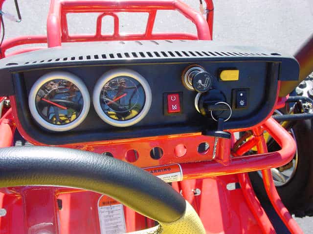 Red sand rail with black steering wheel and dashboard, featuring speed and RPM gauges, headlights switch, and warning sticker.