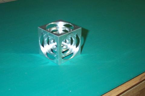 A silver aluminum cube with circular cutouts on each face, sitting on a green surface.
