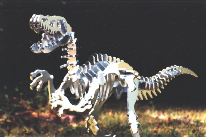 3D laser-cut Velociraptor skeleton model crafted from metal or wood on a blurred outdoor background.
