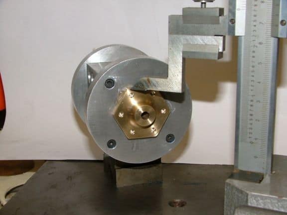 Metal component with hexagonal brass insert on stand, featuring six holes with circular recesses.