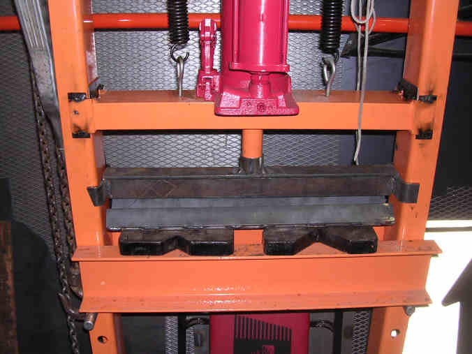 Industrial hydraulic press with red ram, black metal jaws, and orange frame.