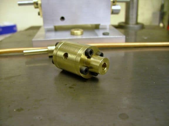 Brass cylindrical object with three holes and thin rod protruding from one end, in an industrial machine shop setting.