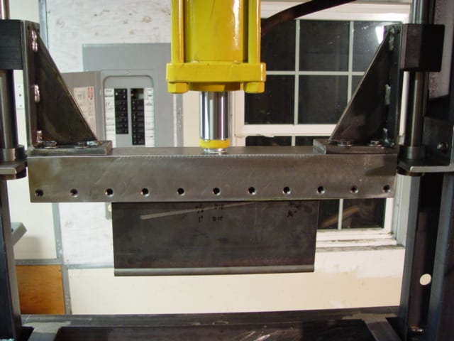 Yellow column metalworking machine with black arms and attached metal component in a workshop background.