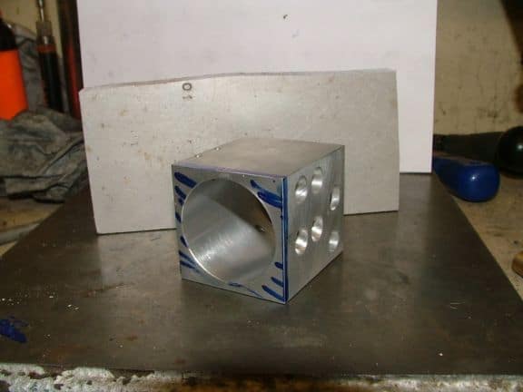 Silver-colored aluminum end cap with square cross-section and circular hole on one side, four holes drilled into it.