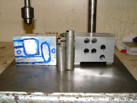 An aluminum block with blue marker markings on a metal surface in front of a drill press or milling machine.