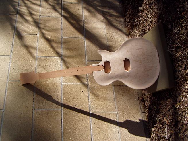 Partially constructed guitar body with light-colored wood back and unattached neck on a concrete floor.