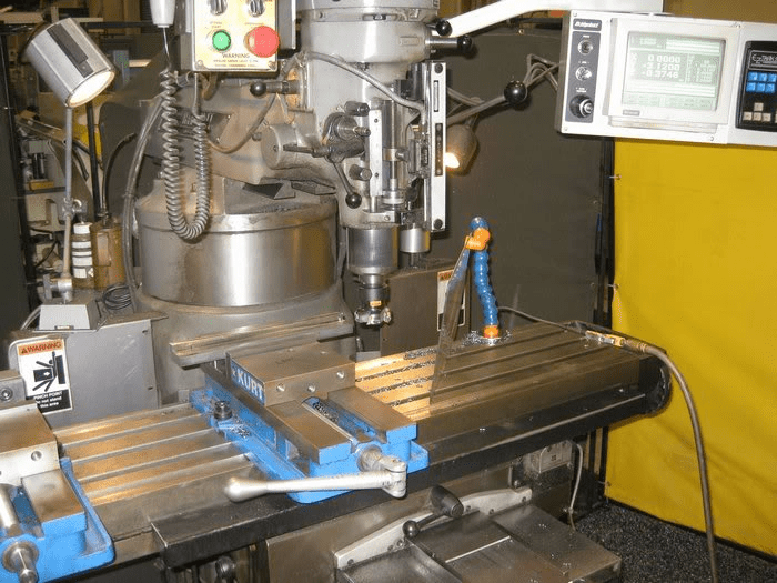 Industrial-grade milling machine with drill press at its center, surrounded by tools and metal components, featuring a CAUTION warning label.